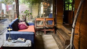 A cozy cabin interior with a blue sofa, a small table with a vase and books, a wooden chair, and a large window with a view of greenery outside. - B&B Hostel (Punta del Este)