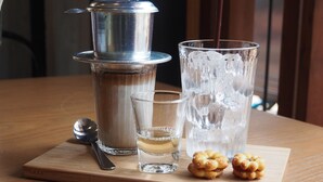 A wooden table with a metal container, two glasses, a spoon, and two small pastries. - Bangkok Story - Hostel (Bangkok)