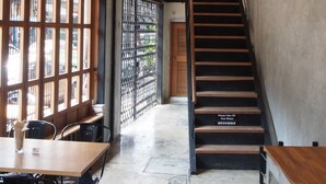 A staircase leading to a second floor, a dining area with tables and chairs, and a view through a window with a metal fence. - Bangkok Story - Hostel (Bangkok)