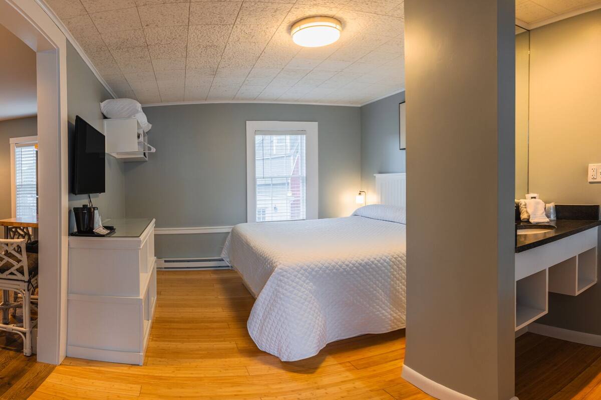 A bedroom with a bed, bedside table, and a small desk with a chair. There is a bathroom with a toilet, sink, and shower.