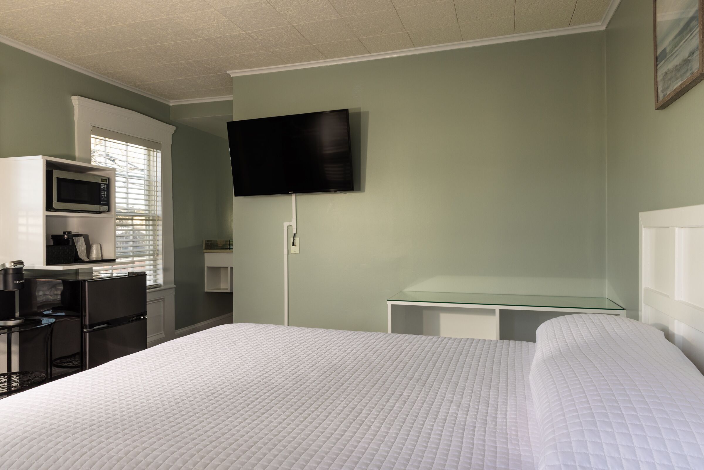 Habitación de hotel con cama, escritorio con silla, microondas, televisión y mesa de vidrio.