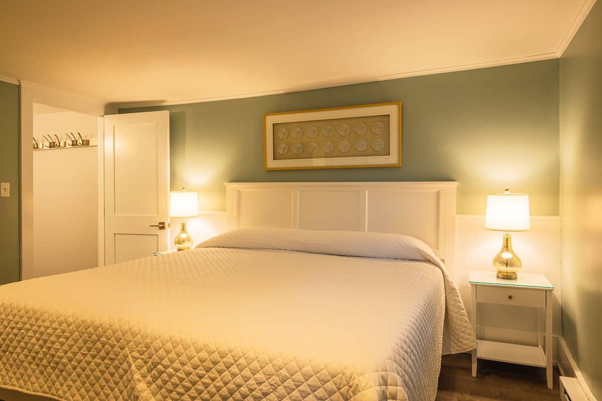 A neatly made bed with a quilted cover, two bedside lamps, a framed picture above the headboard, and a white nightstand.