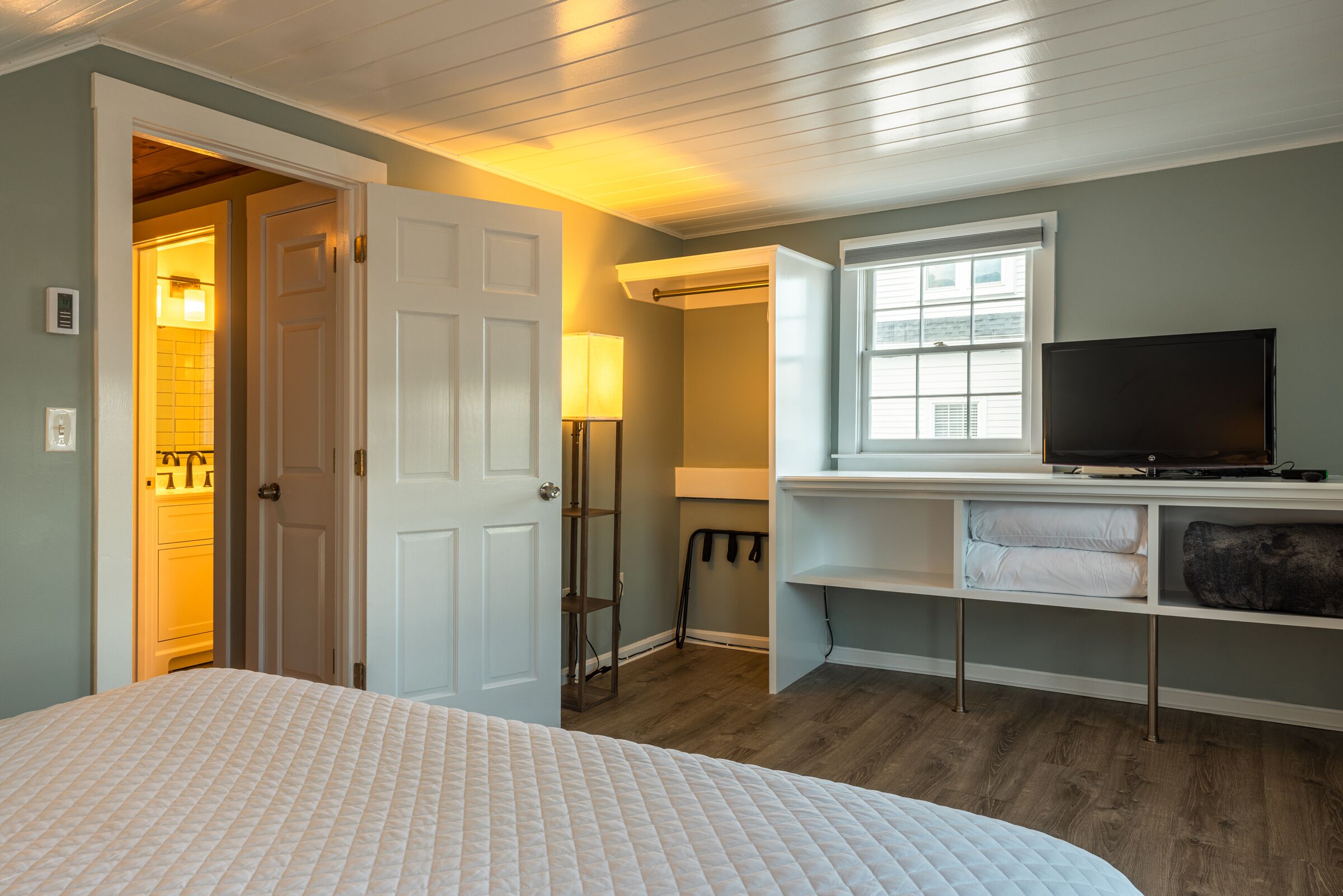 Une chambre à coucher avec un lit, un téléviseur posé sur un meuble, une fenêtre et une porte donnant sur une salle de bain.