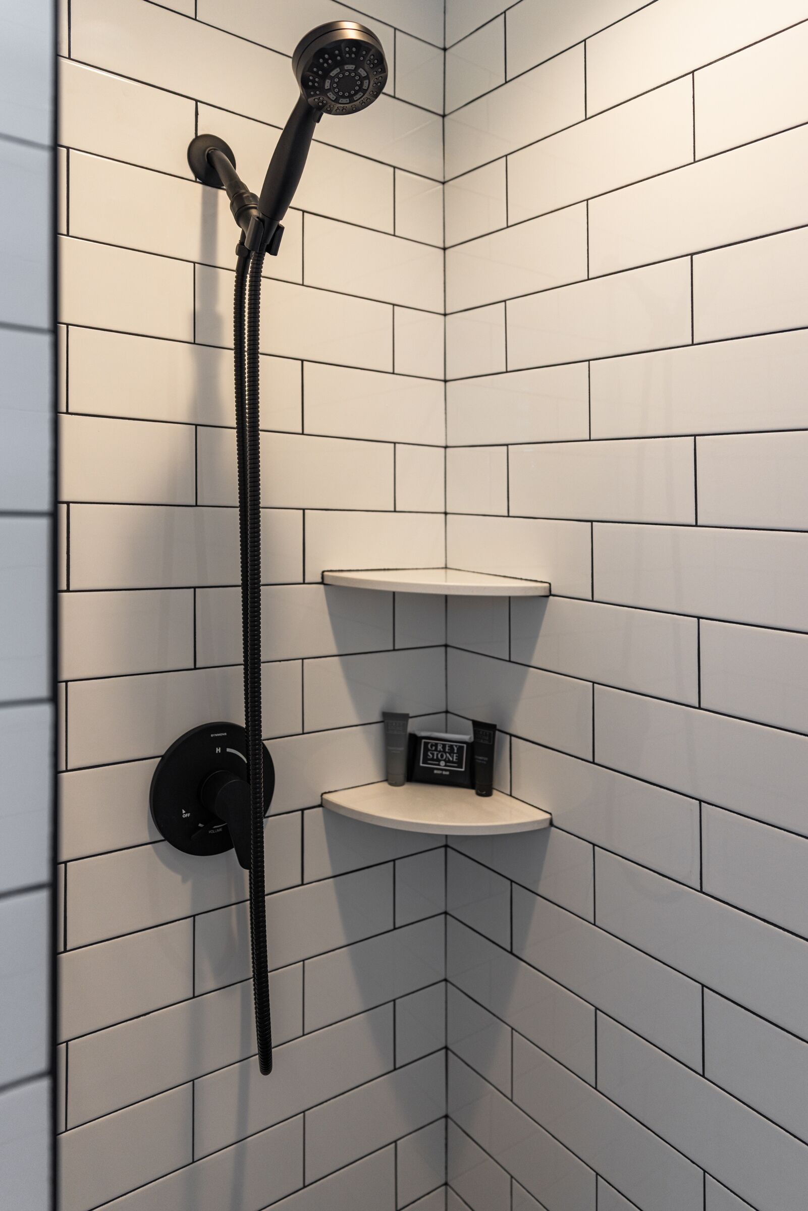 A corner shower with a handheld showerhead, a control panel, and two white shelves.