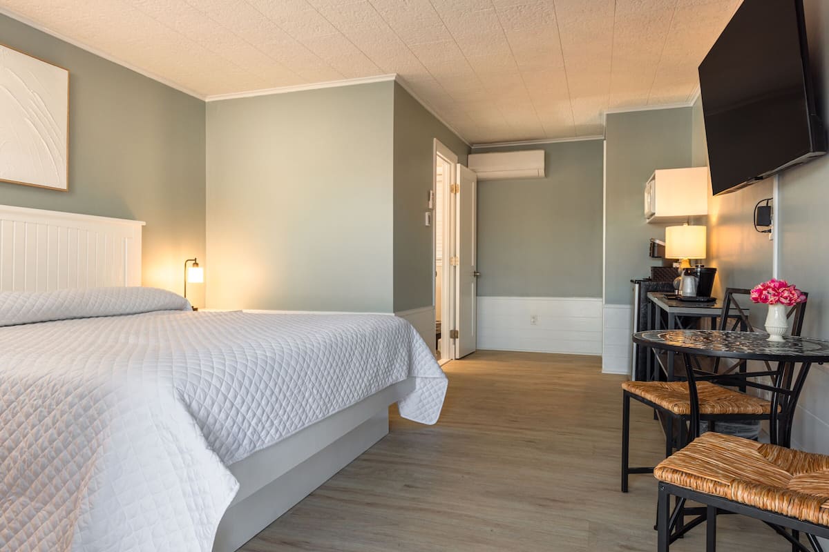 A hotel room with a bed, a dining table with chairs, a TV mounted on the wall, and a door leading to another room.