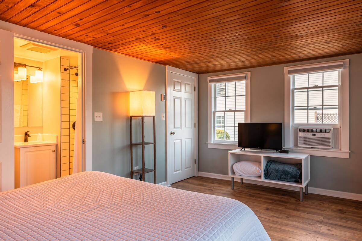 A bedroom with a wooden ceiling, a bed with a white quilt, a television on a white shelf, and a window with blinds.