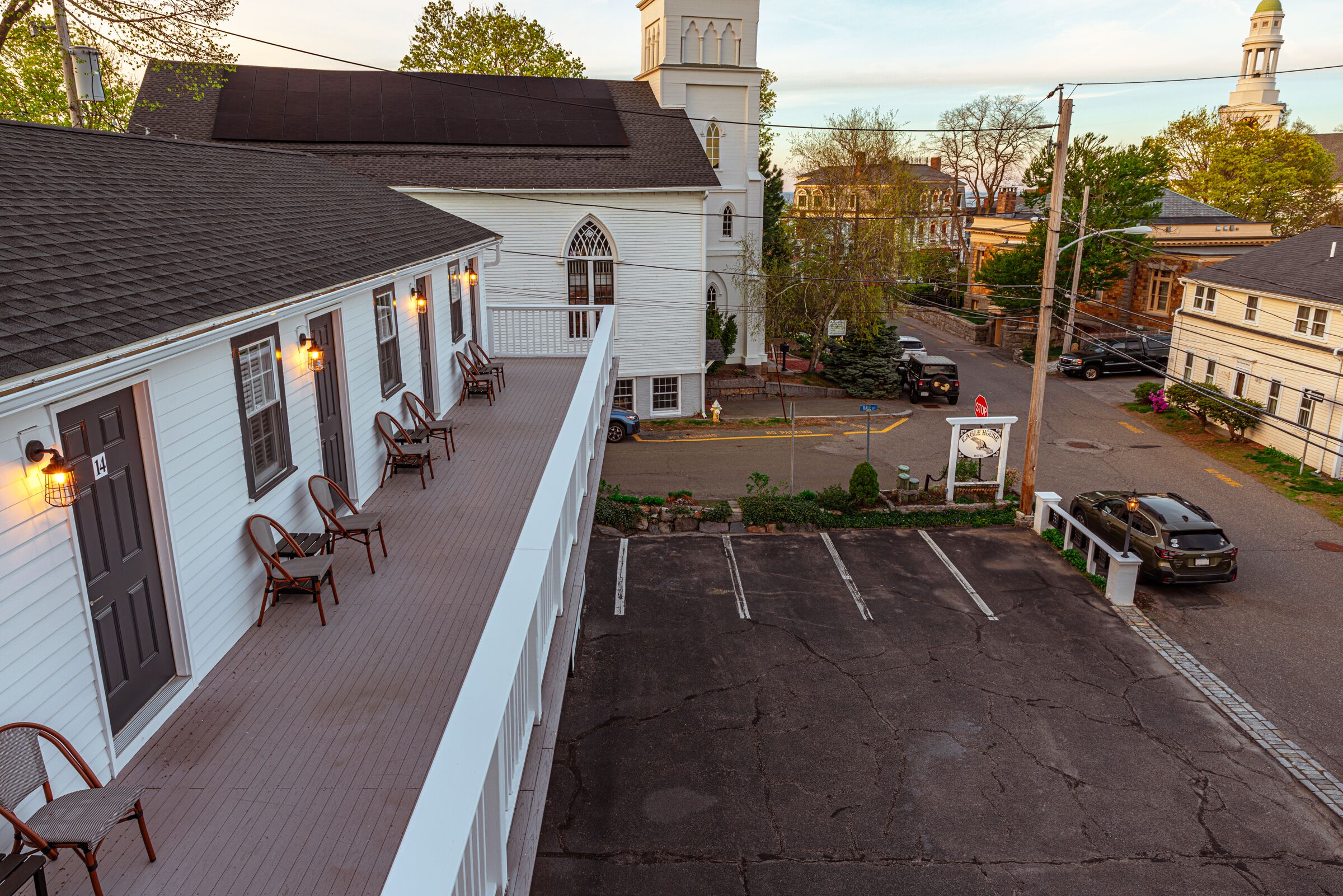 A white building with a porch, a church, and a parking lot.