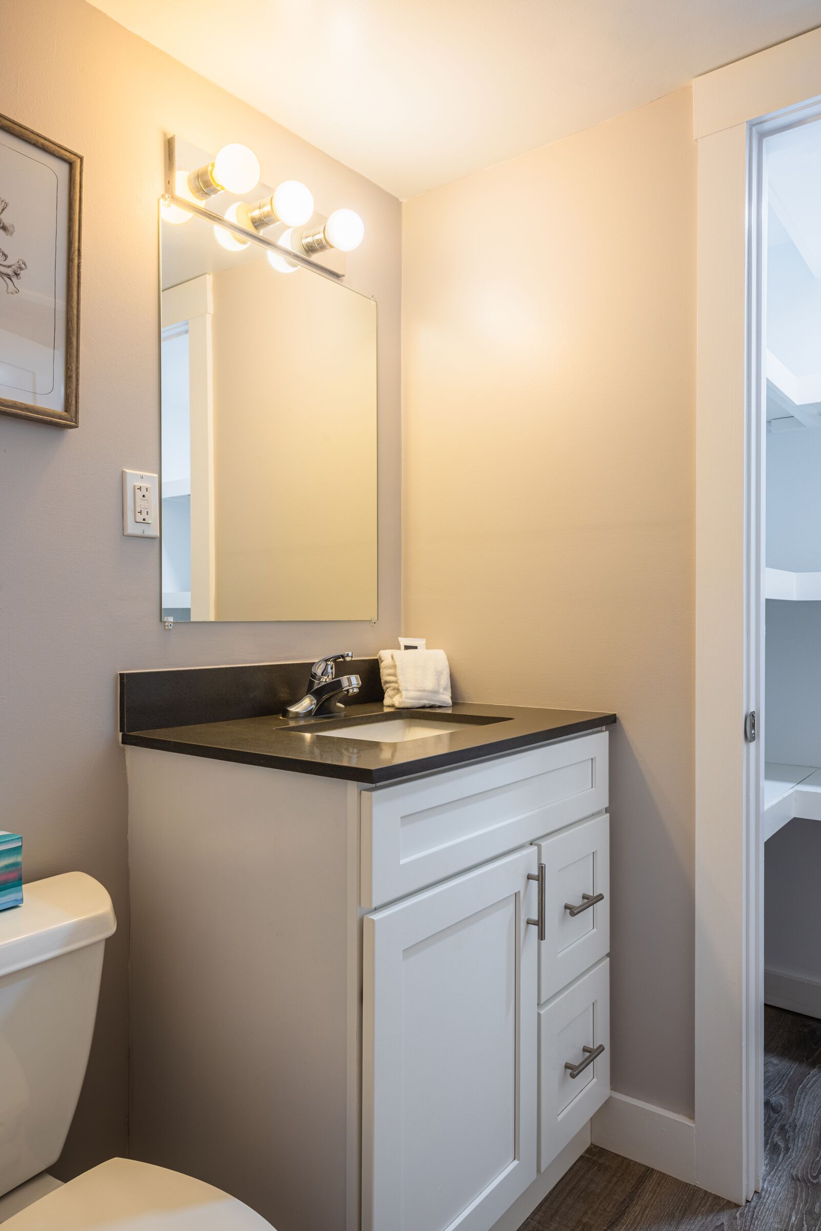 A bathroom with a white toilet, a black sink with a mirror above it, and a white cabinet with drawers. There is a framed picture on the wall and a light switch.