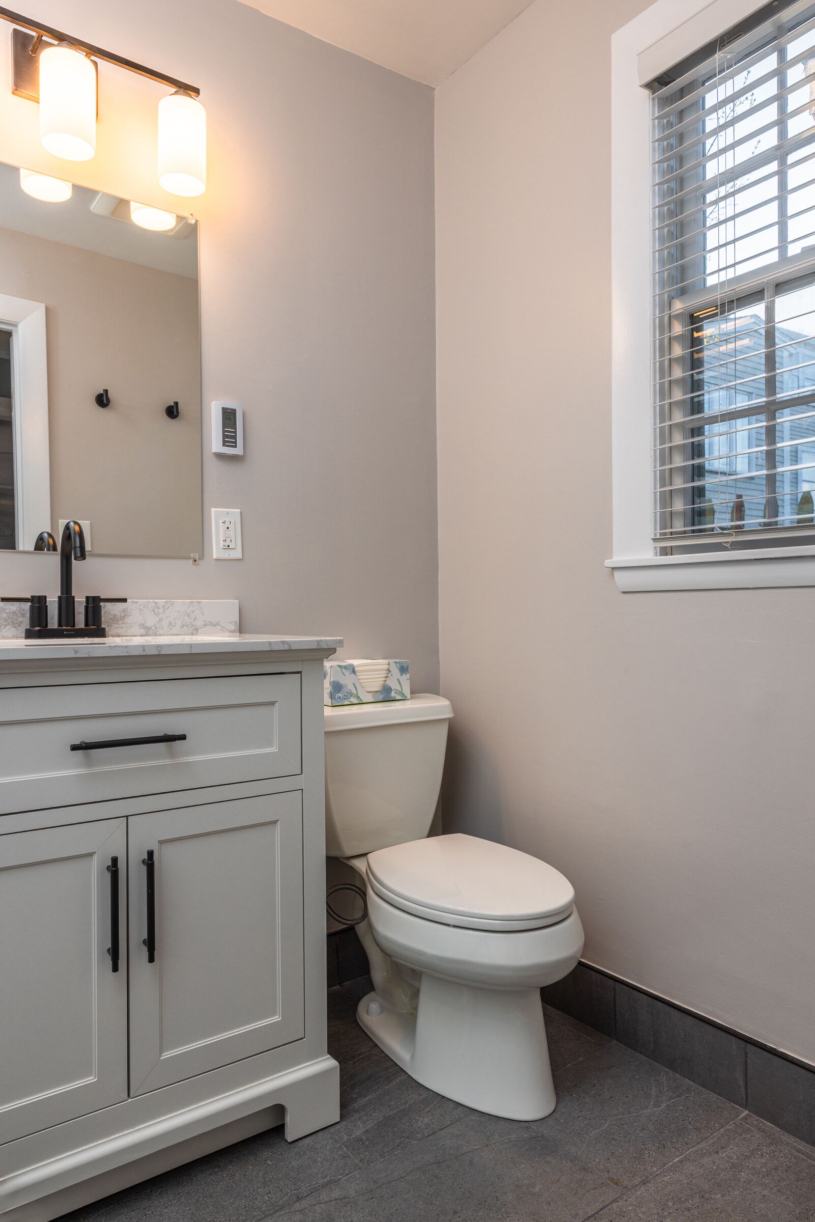 A modern bathroom with a white vanity, a toilet, and a window with blinds.
