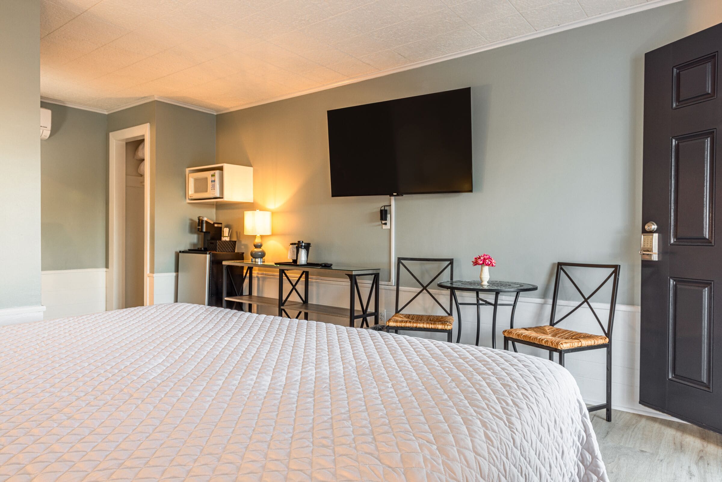 Ein Hotelzimmer mit einem großen Bett, einem Schreibtisch mit Computer, einem flachbildfernseher an der Wand und einer Tür mit Griff.