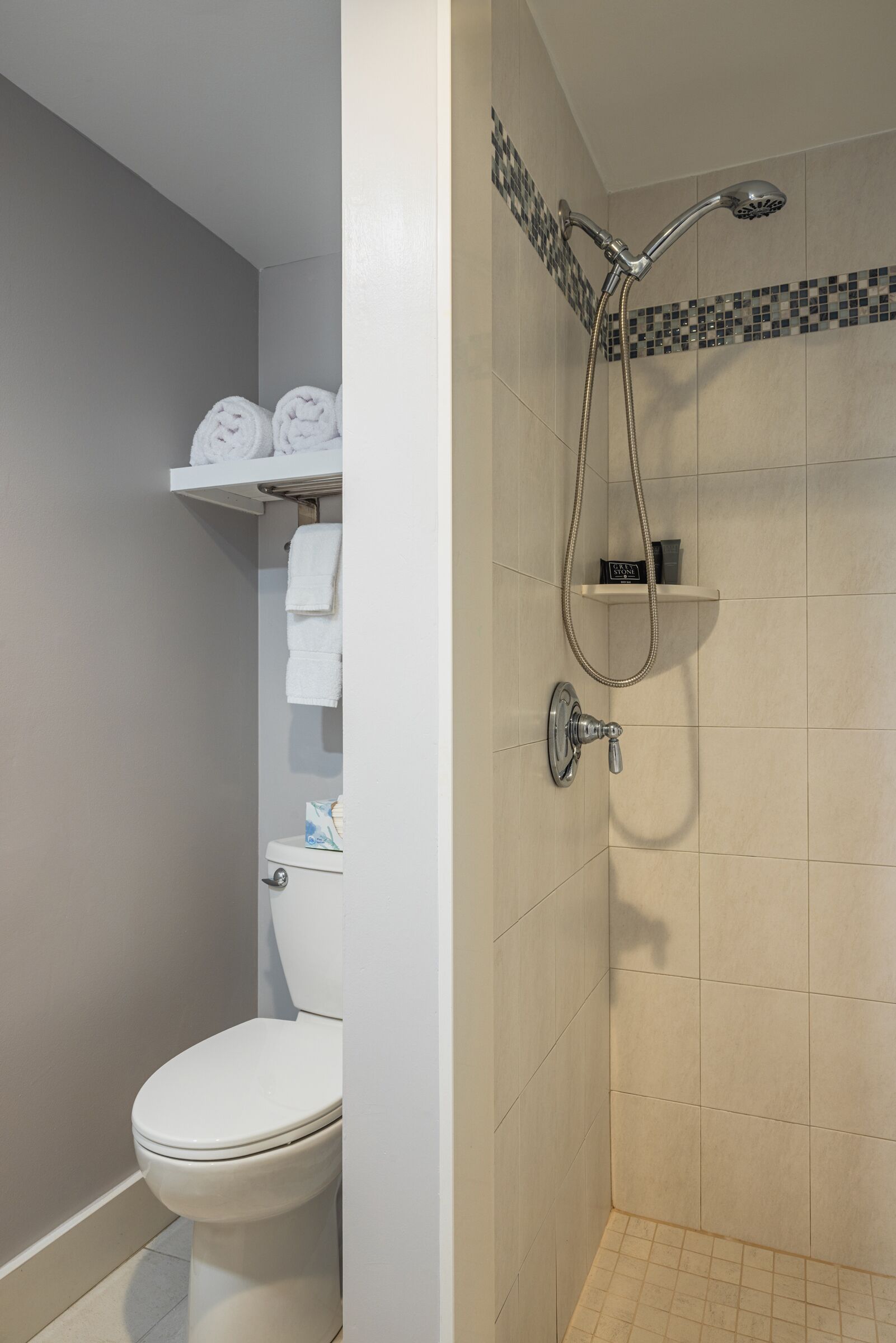 A bathroom with a toilet, a shower with a handheld showerhead, and a shelf with folded towels.