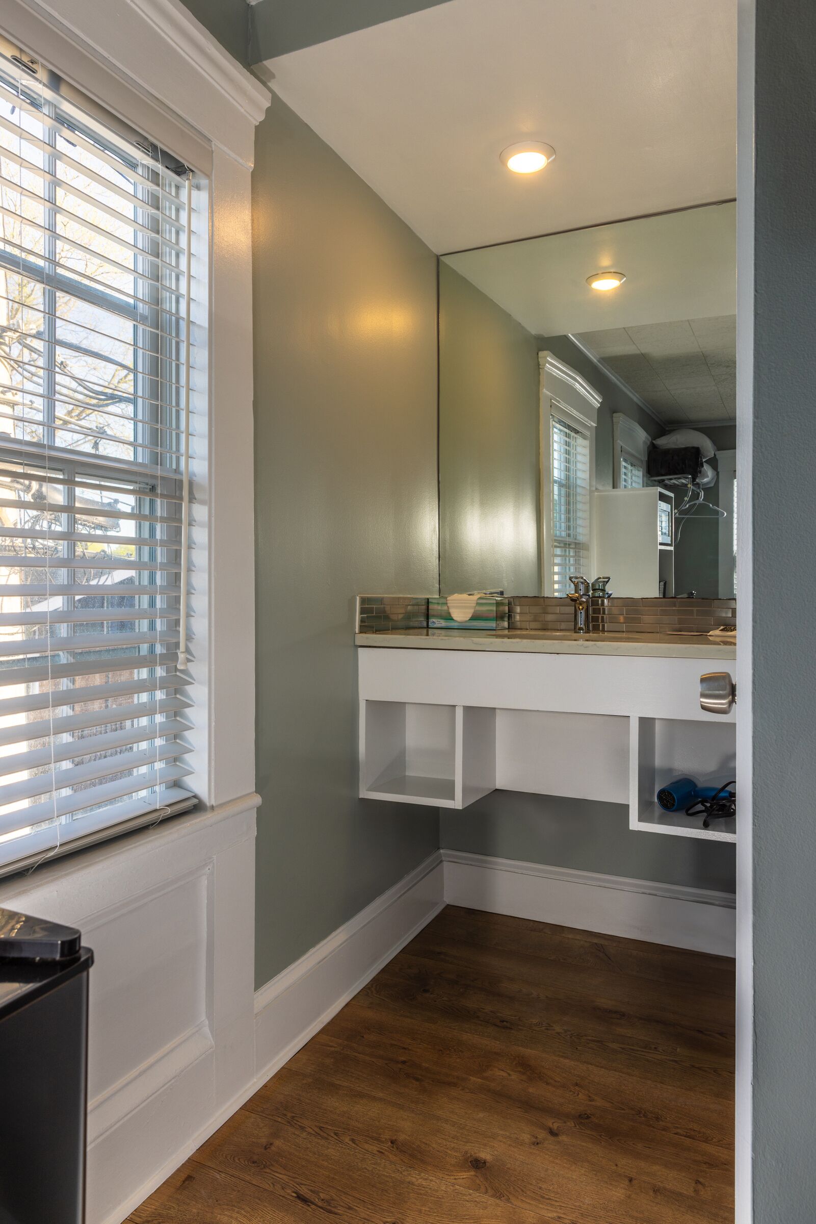 A bathroom with a wooden floor, a white vanity with a mirror, and a window with blinds.