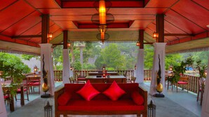 A red sofa with cushions in a covered outdoor seating area with tables and chairs, surrounded by greenery. - E-outfitting Pai Resort (Pai)