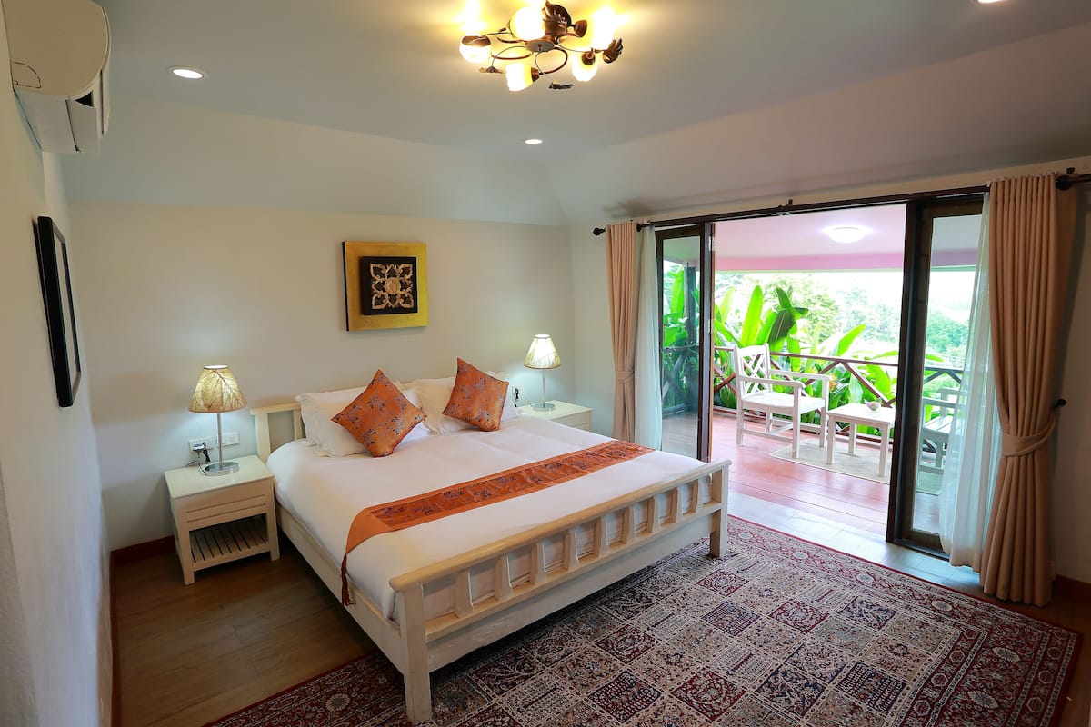 A bedroom with a bed, nightstands, a lamp, a patterned rug, and a view of a balcony with plants.