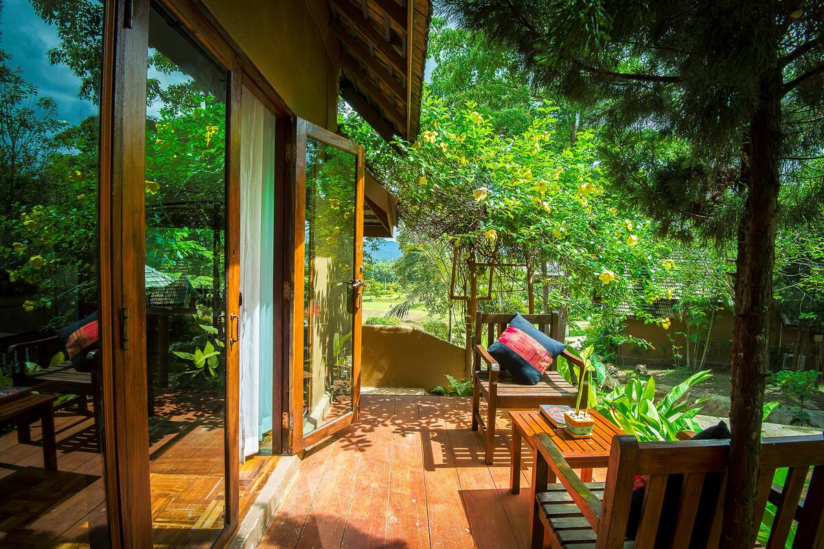 A wooden deck with a bench and a table, surrounded by lush greenery and a view of a garden.