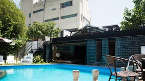 A swimming pool with stone pillars, wicker chairs, and a building with a blue-tiled exterior in the background. - The Style Capsule Guesthouse - Hostel (Daegu)