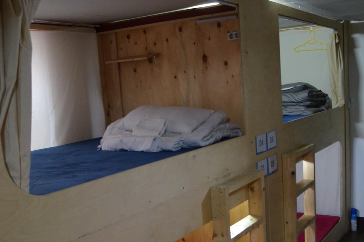 A bunk bed room with a wooden bunk bed, a lower bunk with a red mattress, and a small shelf unit.