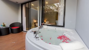 A modern bathroom with a large bathtub, a shower, and a toilet. There is a window with curtains, a small table with a red cushion, and a wicker chair.