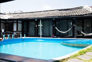 Una piscina con techo de baldosas y puertas de cristal, rodeada de mobiliario exterior y una hamaca.