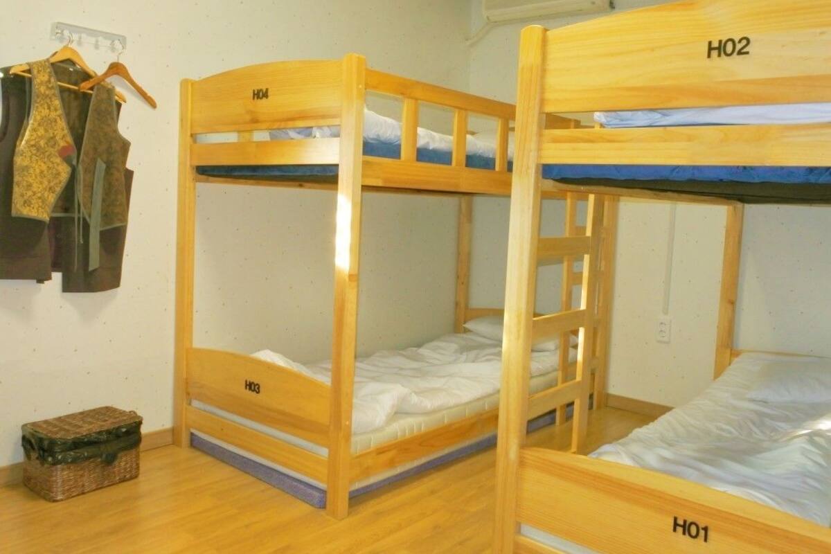 A room with three bunk beds labeled H01, H02, and H03. There is a wooden floor, a wall-mounted heater, and a small basket on the floor.