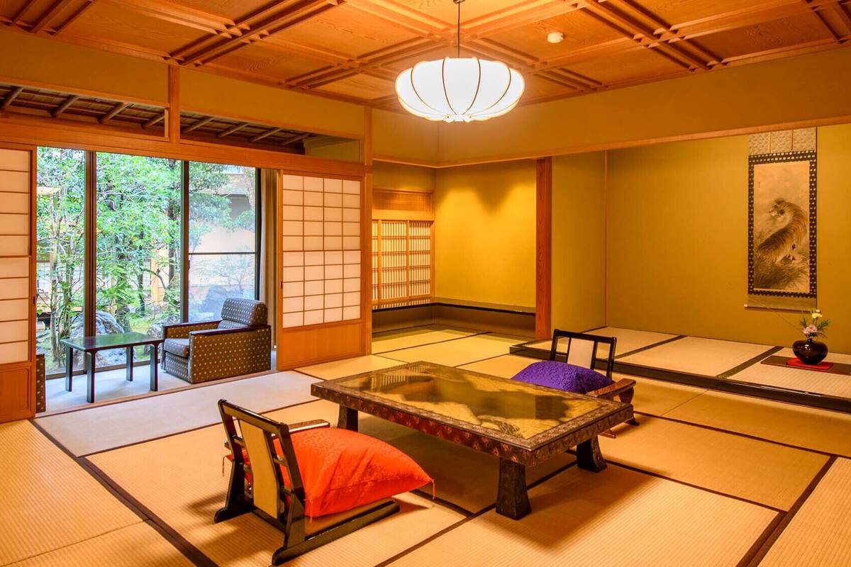 A traditional Japanese room with tatami flooring, a low table, a cushioned chair, and sliding doors leading to a garden.