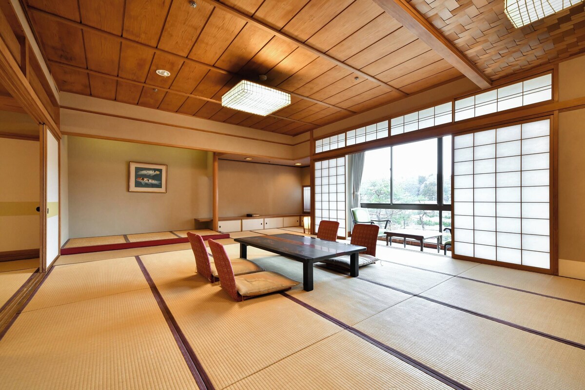 A traditional tatami mat room with a low table and chairs, large windows, and a ceiling with wooden beams.
