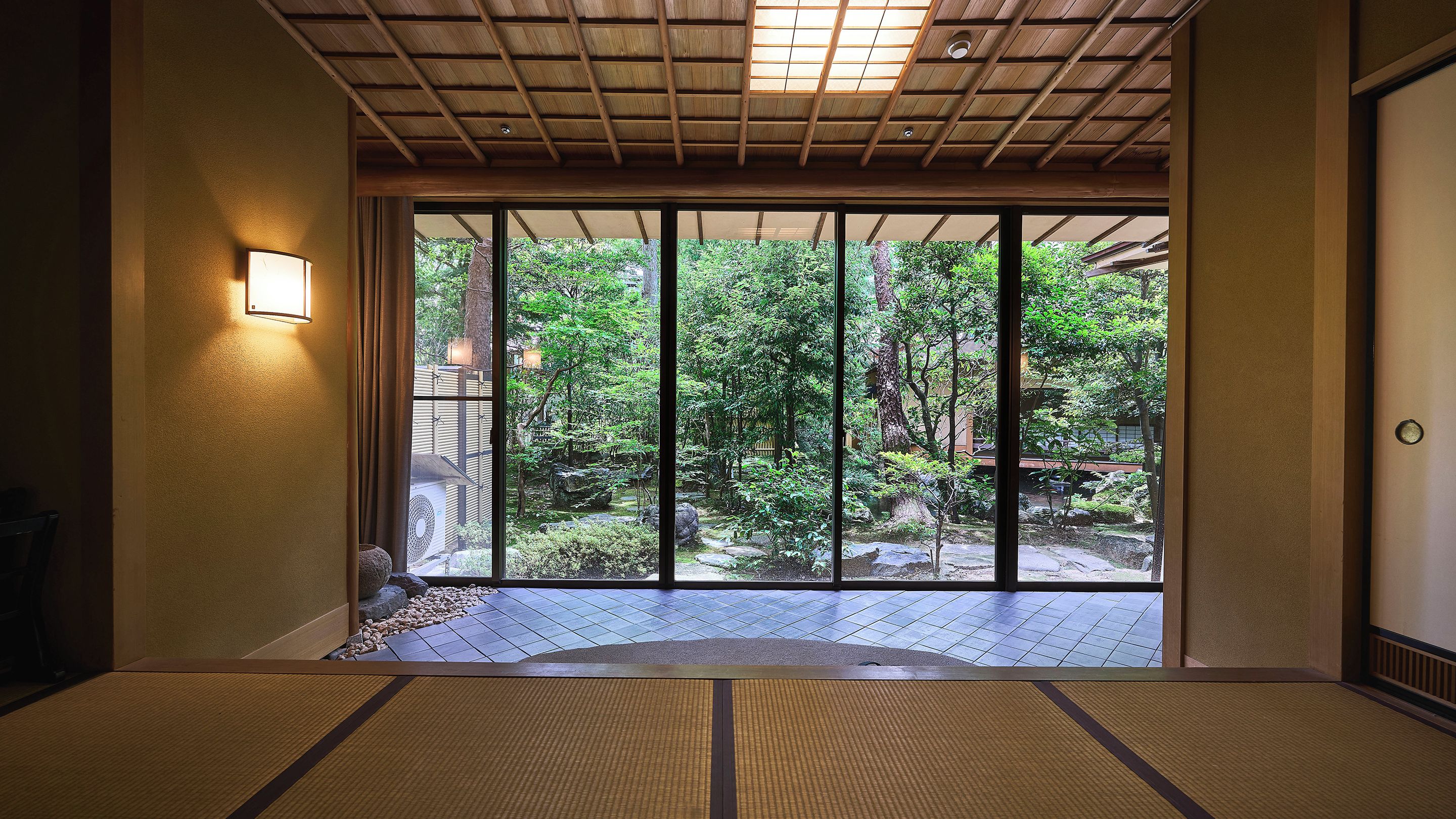 A traditional Japanese room with tatami flooring, a sliding door leading to a garden, and a ceiling with wooden beams.
