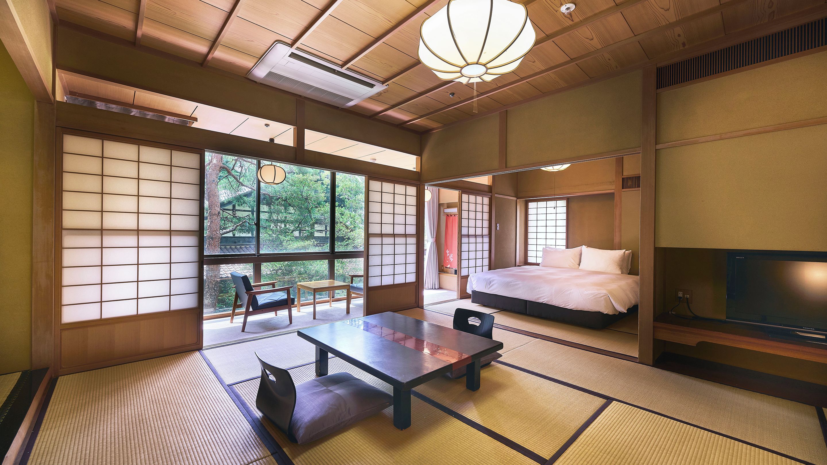 Une pièce de style japonais traditionnel, avec un sol recouvert de tatamis, un lit, une télévision et une vue sur la verdure environnante.
