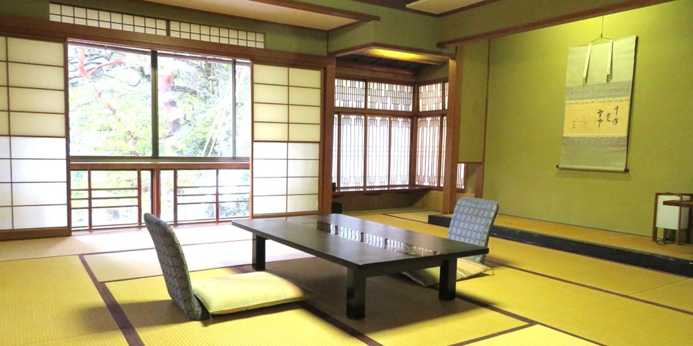 A tatami mat room with a low table and chairs, large windows with shoji screens, and a wall with calligraphy.
