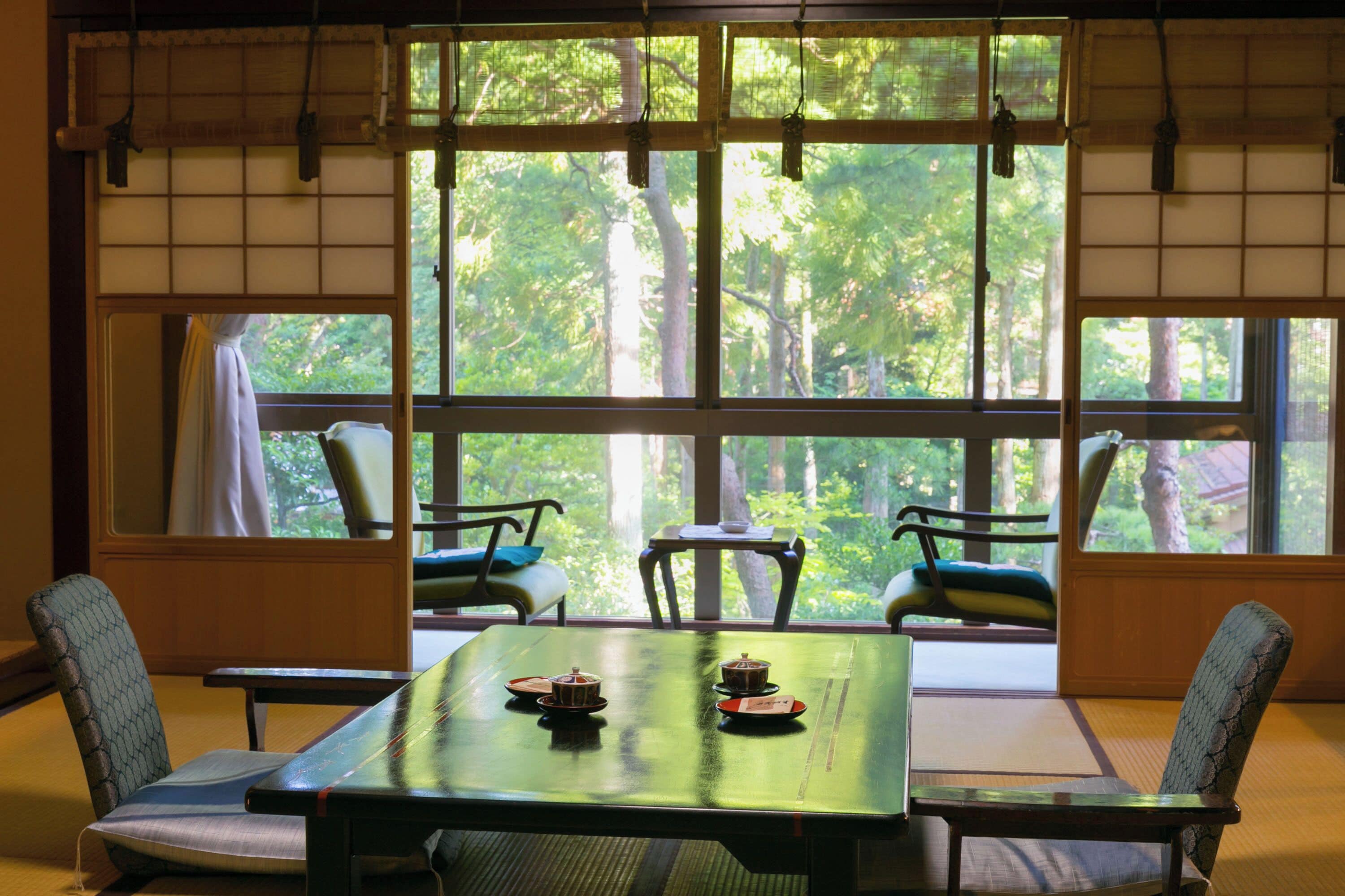 Une pièce de style japonais traditionnel, avec un sol recouvert de tatamis, une petite table basse agrémentée de services à thé et de grandes fenêtres donnant sur une forêt.