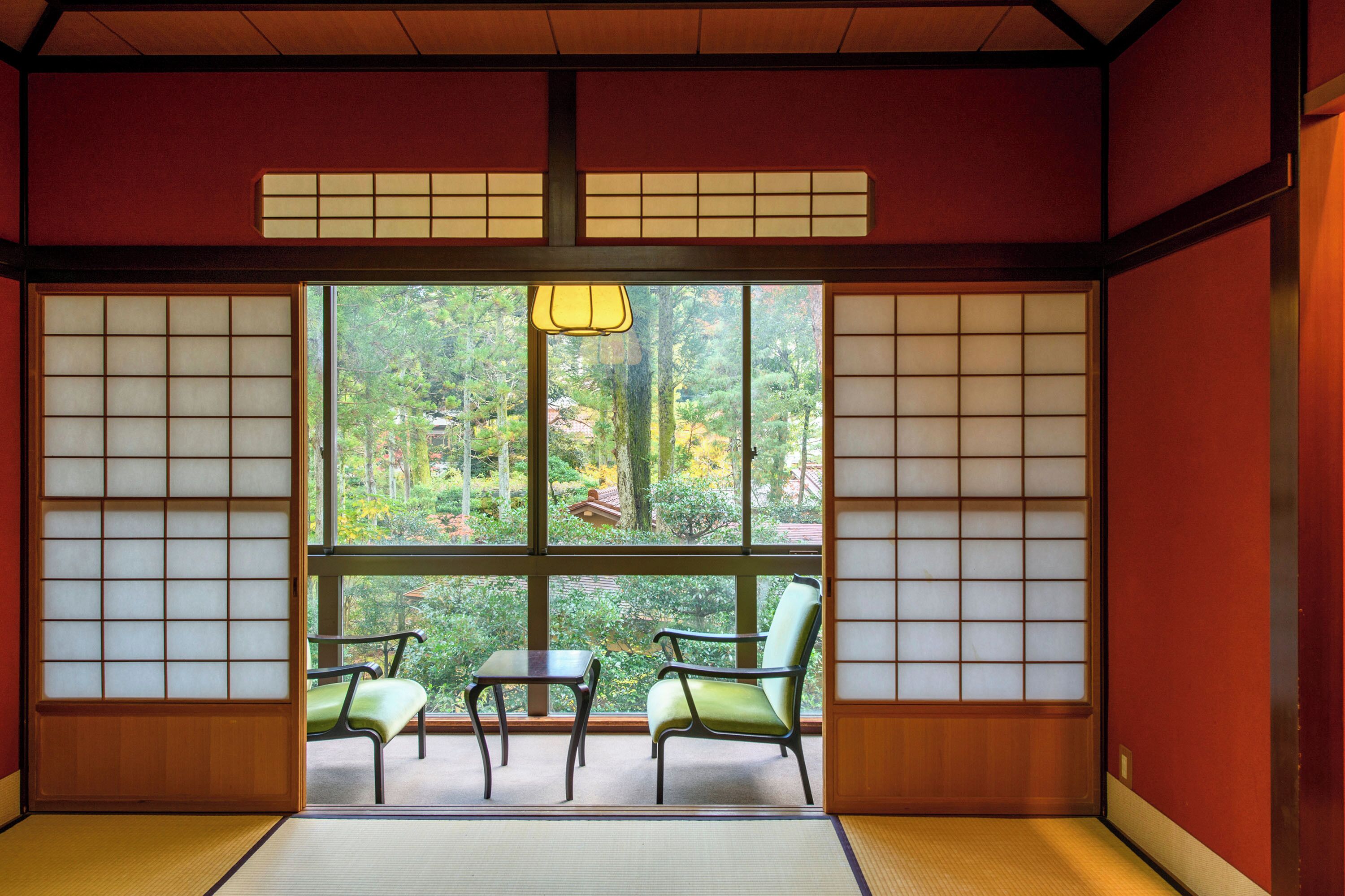 A traditional Japanese room with tatami flooring, a low table, two chairs, and a view of a forest through shoji screens.