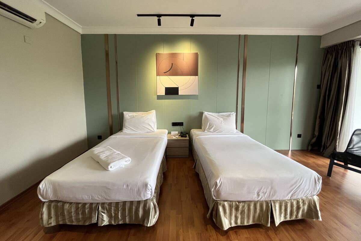 A hotel room with two single beds, a nightstand with a lamp, a wall-mounted artwork, and a wooden floor.