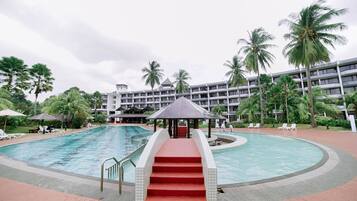 A swimming pool with red steps leading to a gazebo, surrounded by palm trees and a multi-story building.