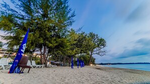 A beach with white lounge chairs, a blue umbrella, and a row of blue flags. - Tunamaya Beach & Spa Resort – Desaru (Bandar Penawar)