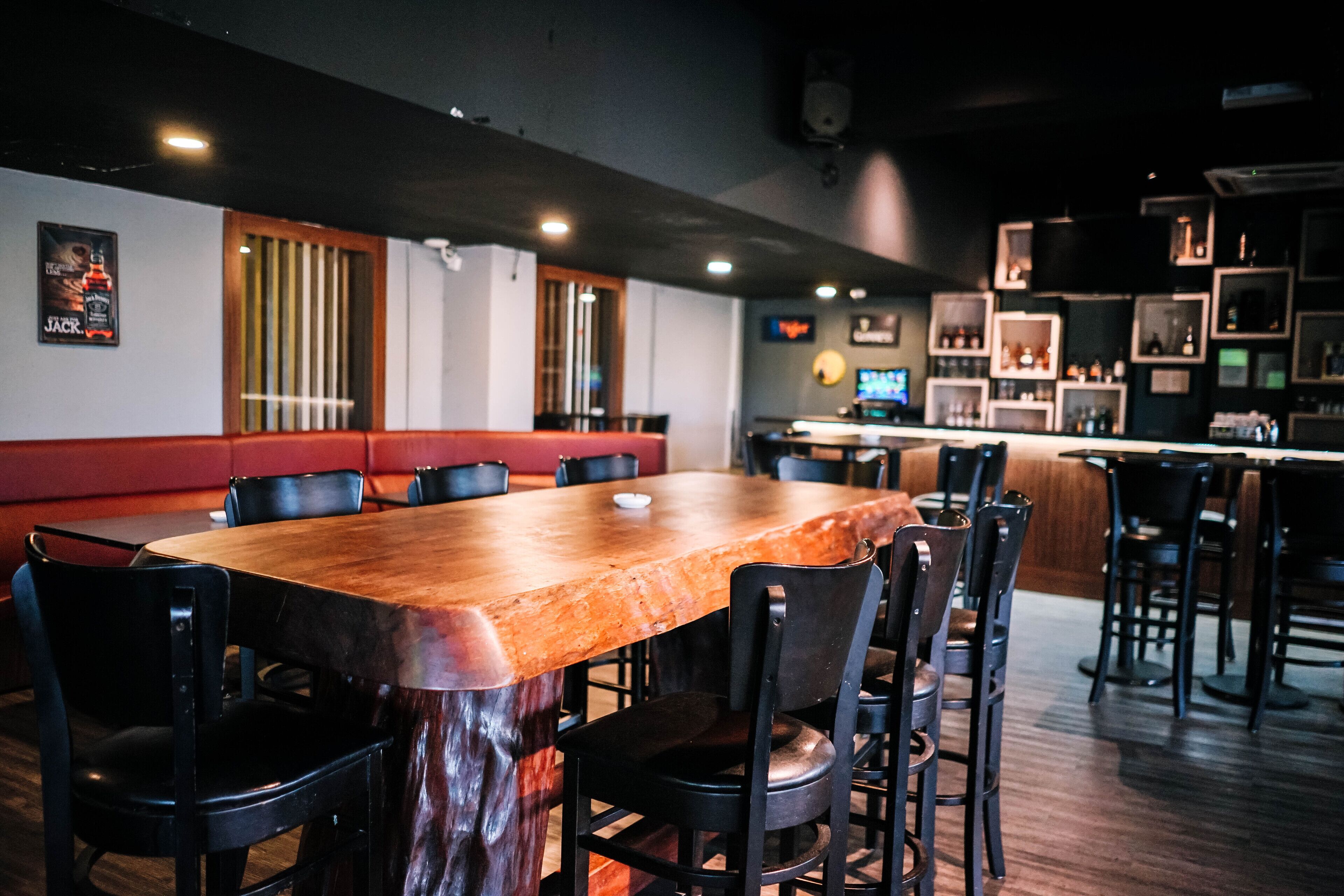 A restaurant with a long wooden table, black chairs, and a bar area with shelves of bottles.
