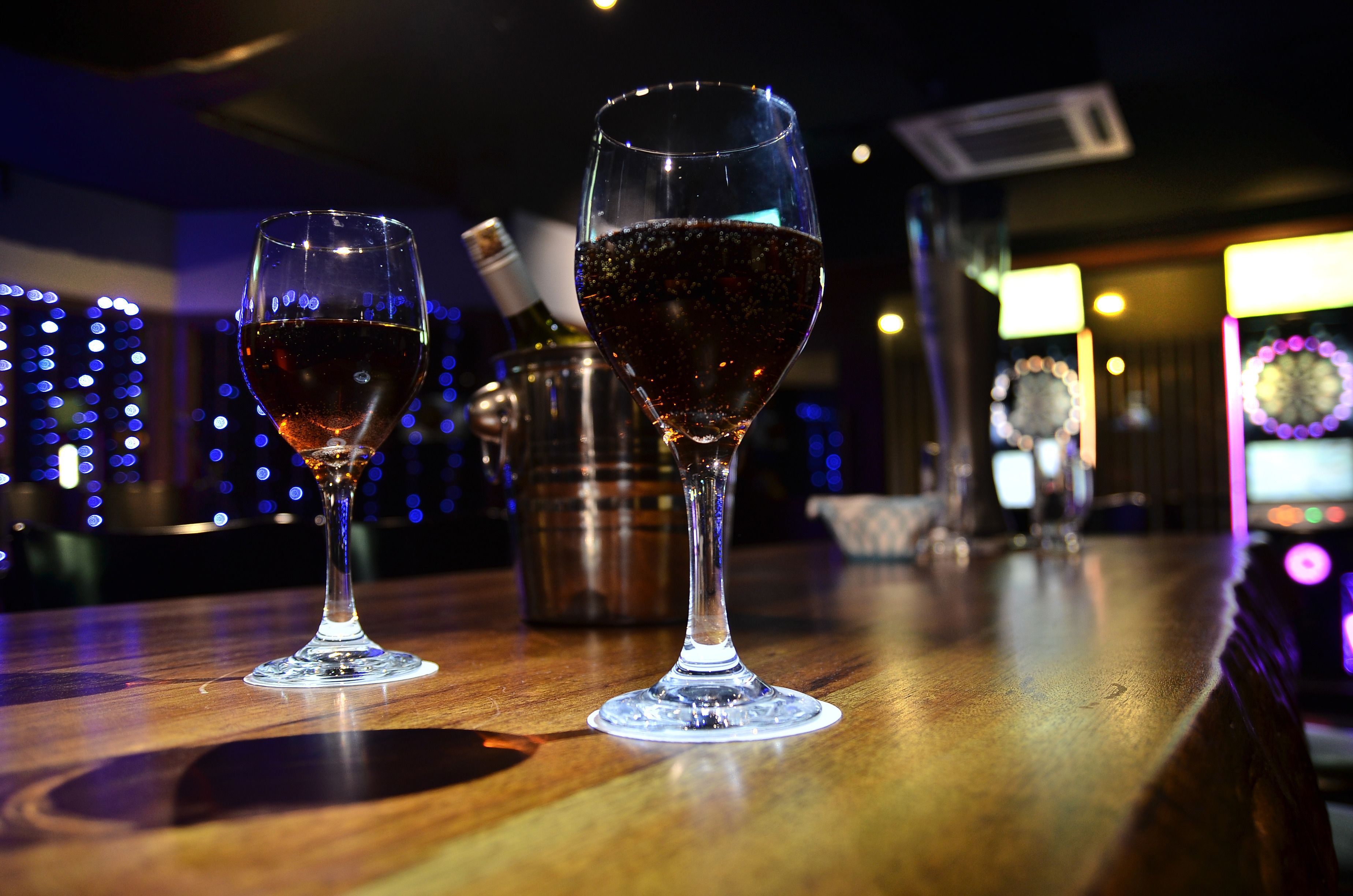Duas taças de vinho sobre um balcão de madeira, com fundo desfocado de luzes e um relógio.