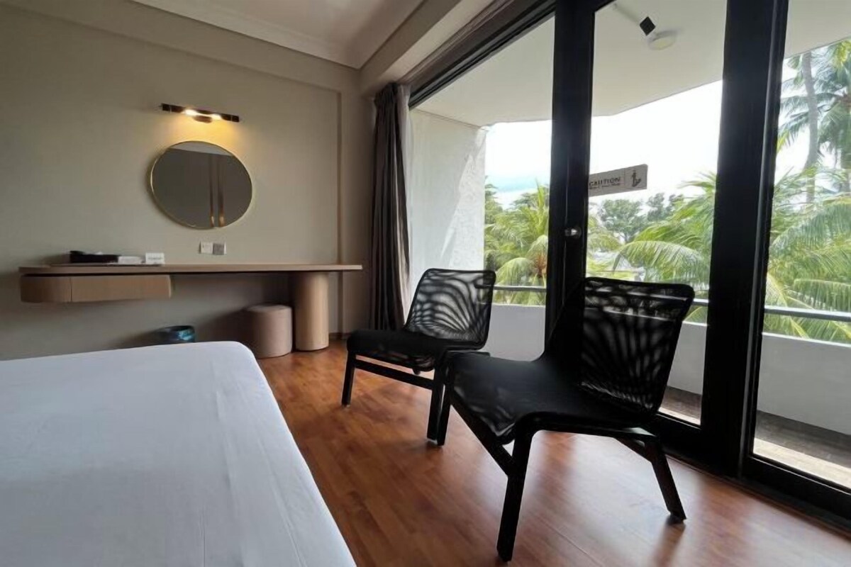 Ein Hotelzimmer mit Bett, Stuhl, Schreibtisch, Spiegel und einem Balkon mit Blick auf Palmen.