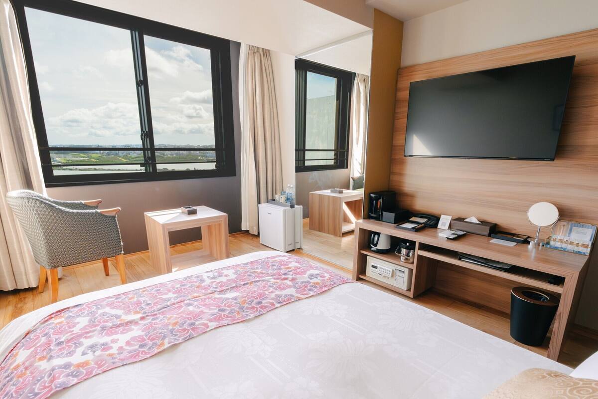 A hotel room with a bed, a desk, a chair, a TV, and a view of the city.