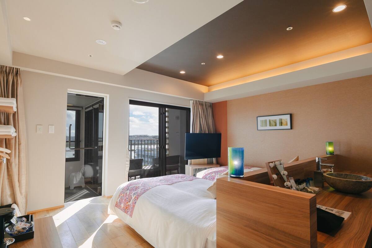 A modern hotel room with a large bed, a wooden desk, a TV, and a view of the city through the balcony doors.