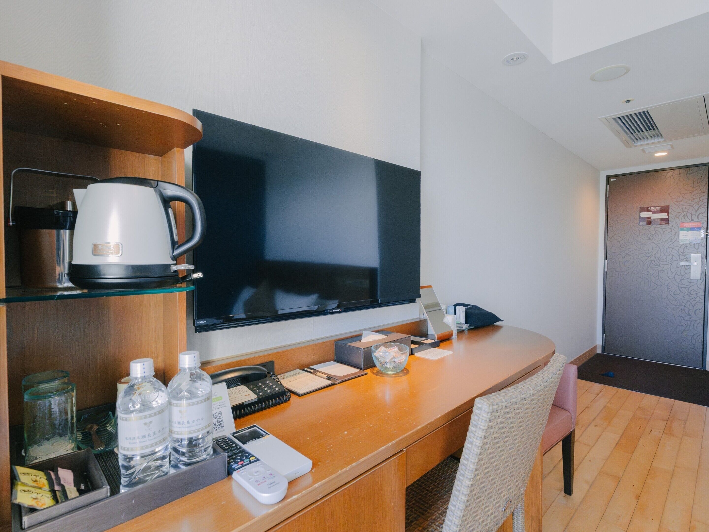 A hotel room with a wooden desk, a flat-screen TV, a coffee maker, a refrigerator, and a wicker chair.