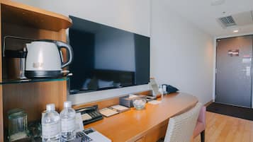 A hotel room with a wooden desk, a flat-screen TV, a coffee maker, a refrigerator, and a wicker chair.