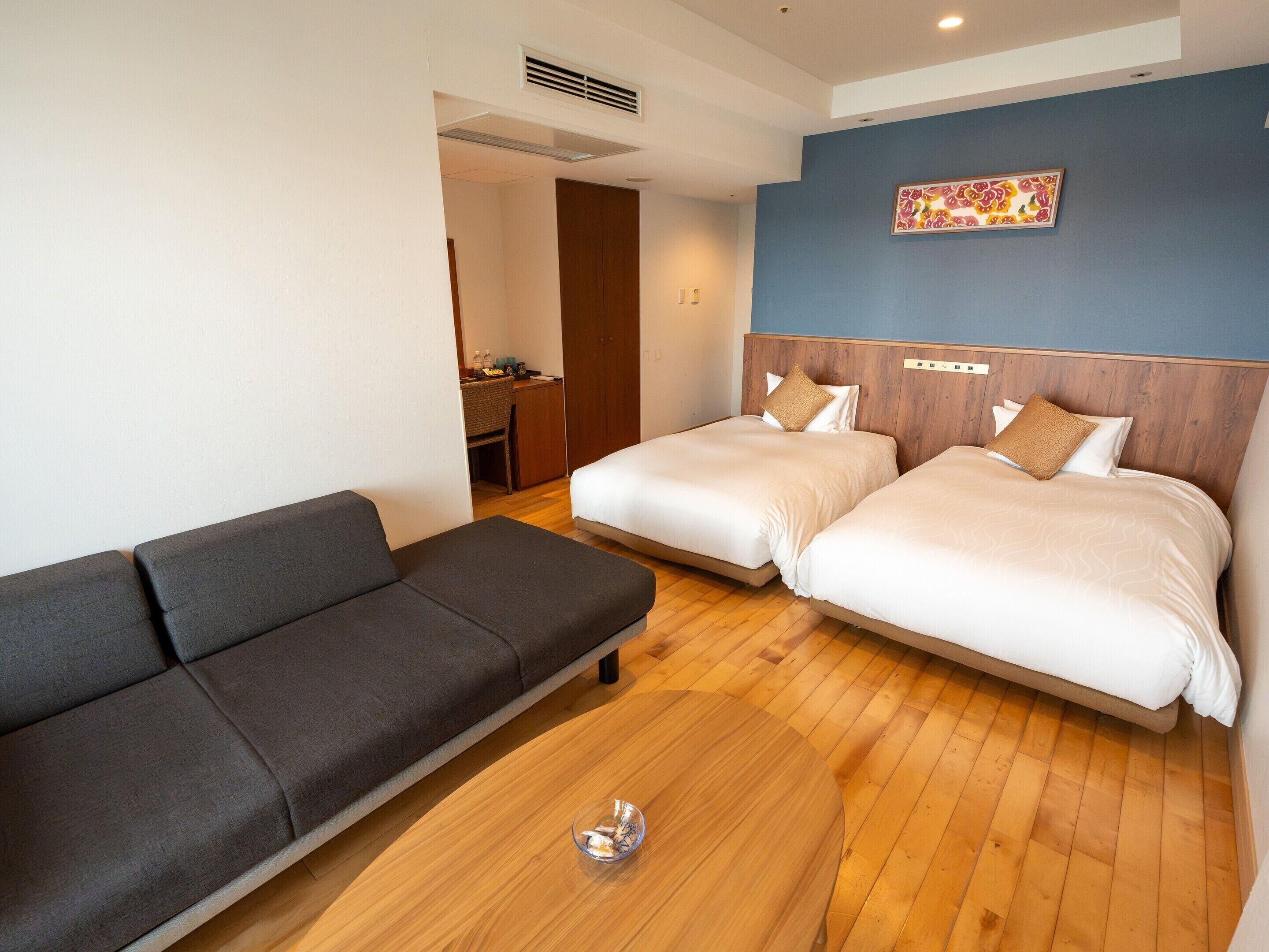 A hotel room with two beds, a sofa, a wooden floor, and a wall-mounted artwork.