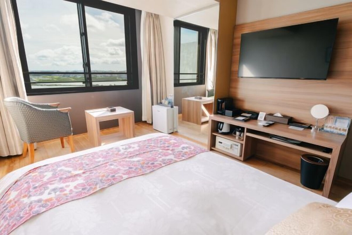Habitación de hotel con cama, escritorio, silla, televisor y vistas al exterior.