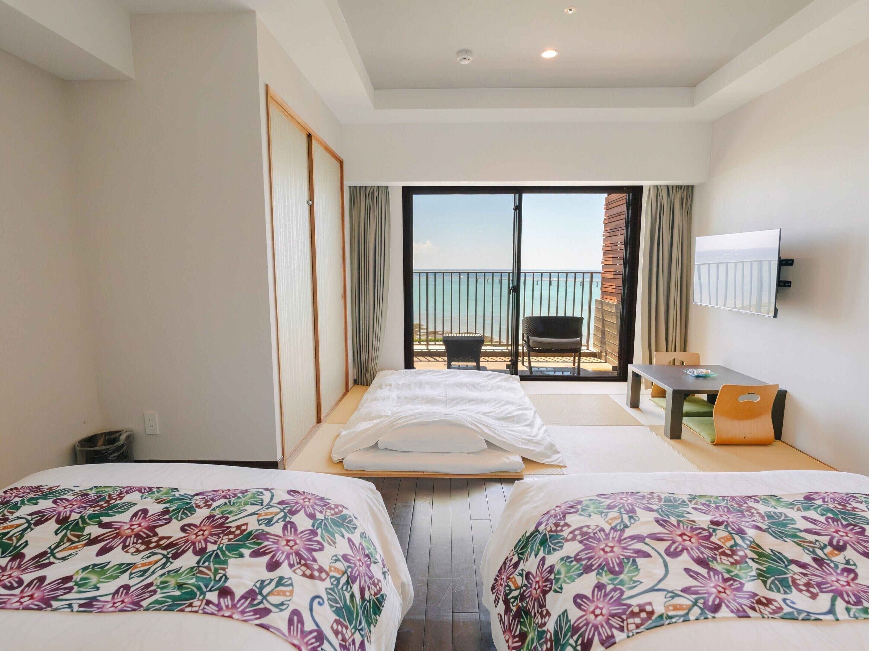 A hotel room with two beds, a desk, a TV, and a balcony with a view of the sea.