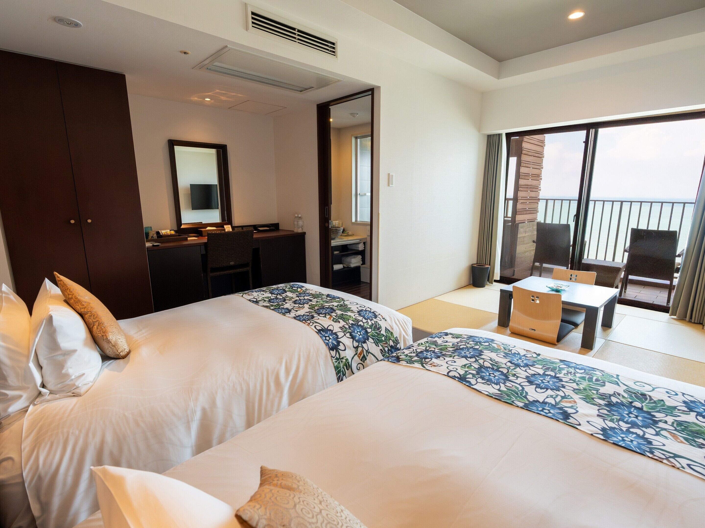 A hotel room with two beds, a desk, a chair, and a balcony with a view.