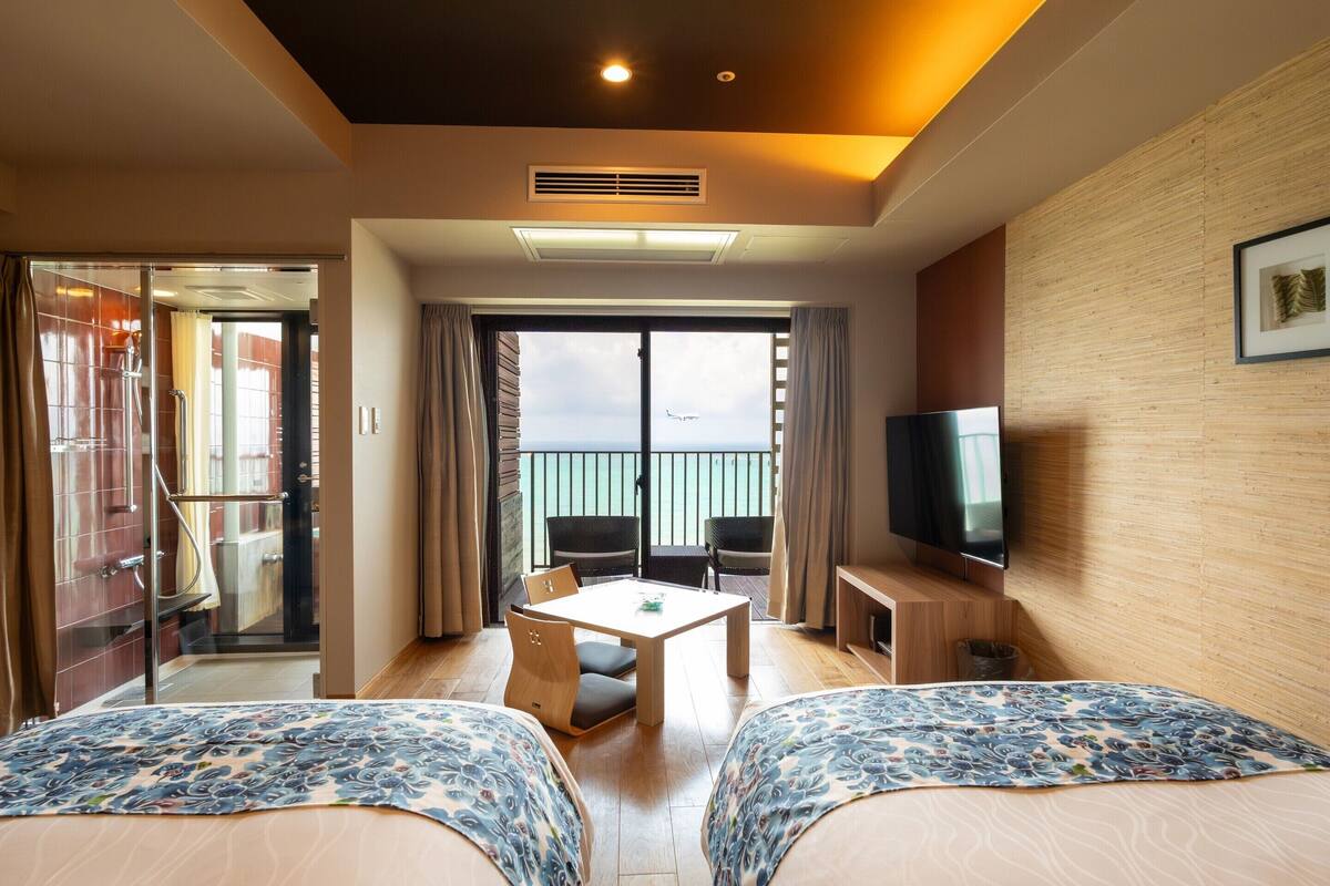 A hotel room with two beds, a TV, a balcony with a view, and a table with chairs.