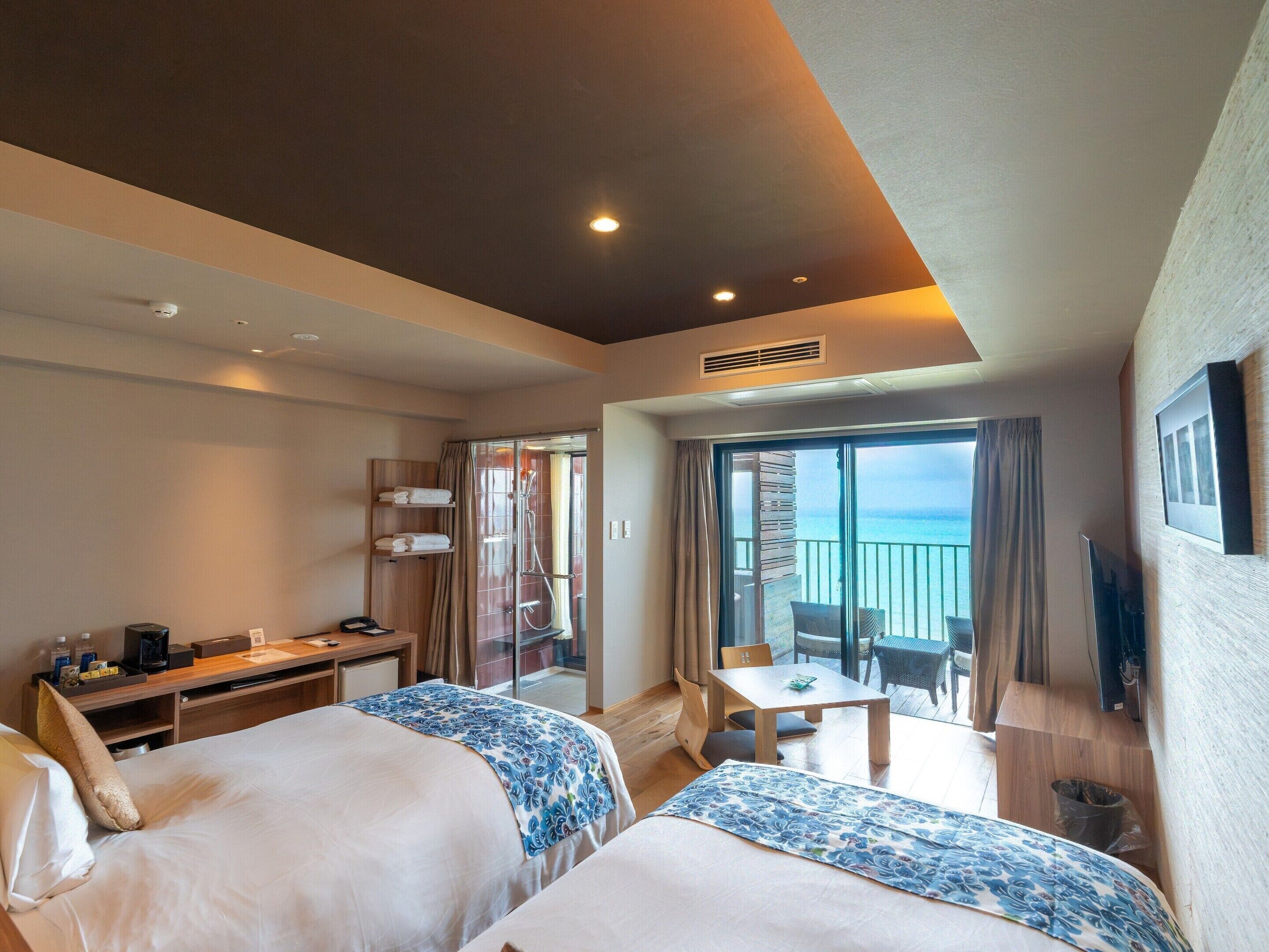 Une chambre d’hôtel avec deux lits, un bureau, une chaise, une petite table et un balcon avec vue sur la mer.