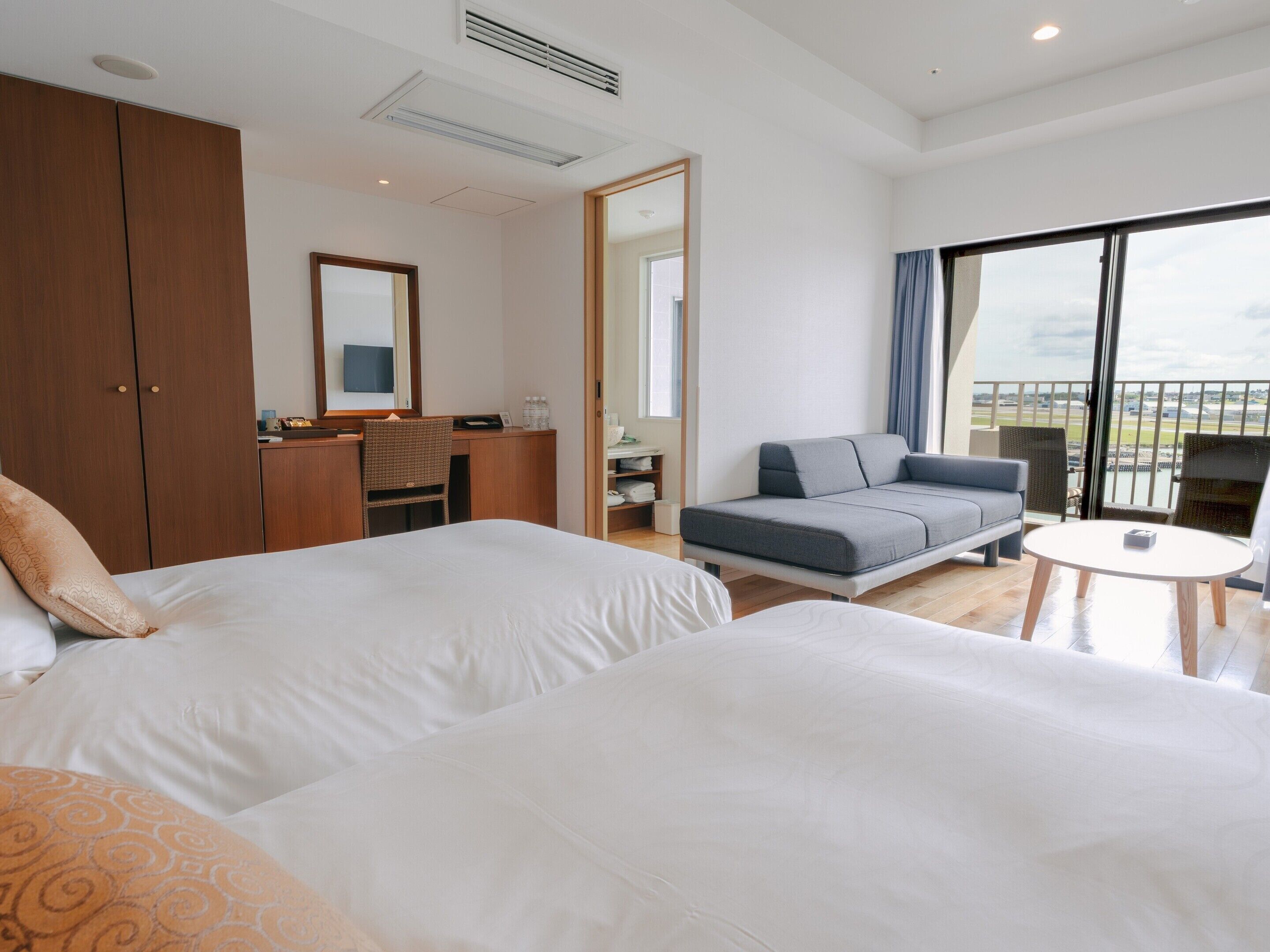 A hotel room with two beds, a sofa, a desk, and a balcony with a view.