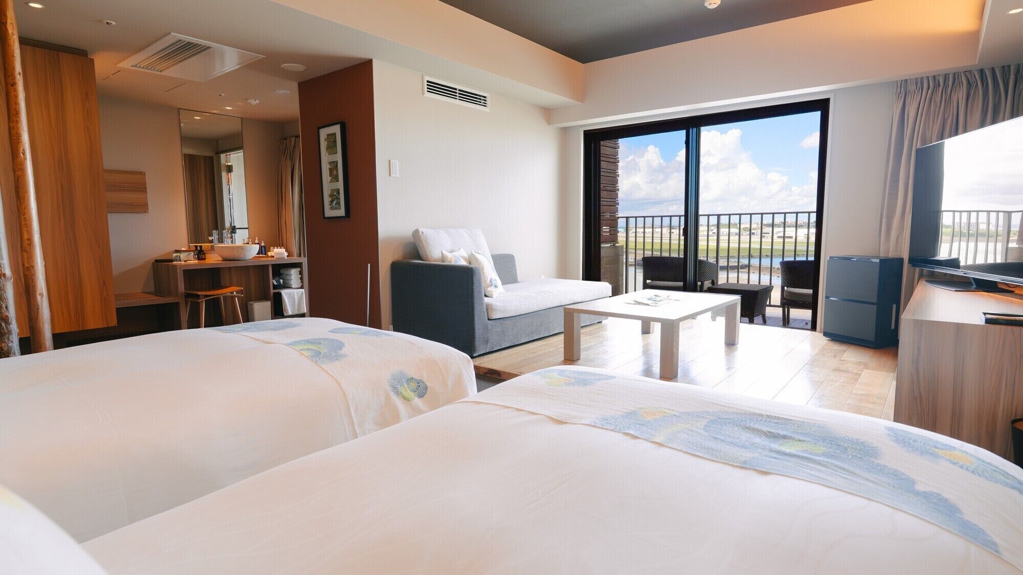 A hotel room with two beds, a sofa, a small table, a TV, and a balcony with a view.