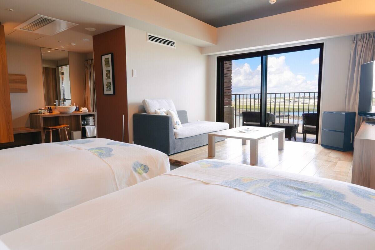 A hotel room with two beds, a sofa, a small table, a TV, and a balcony with a view.